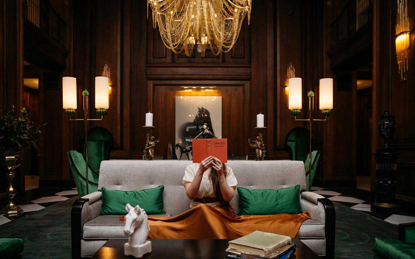 a woman reading a book in a hotel lobby with rich dark wood walls and a golden chandelier